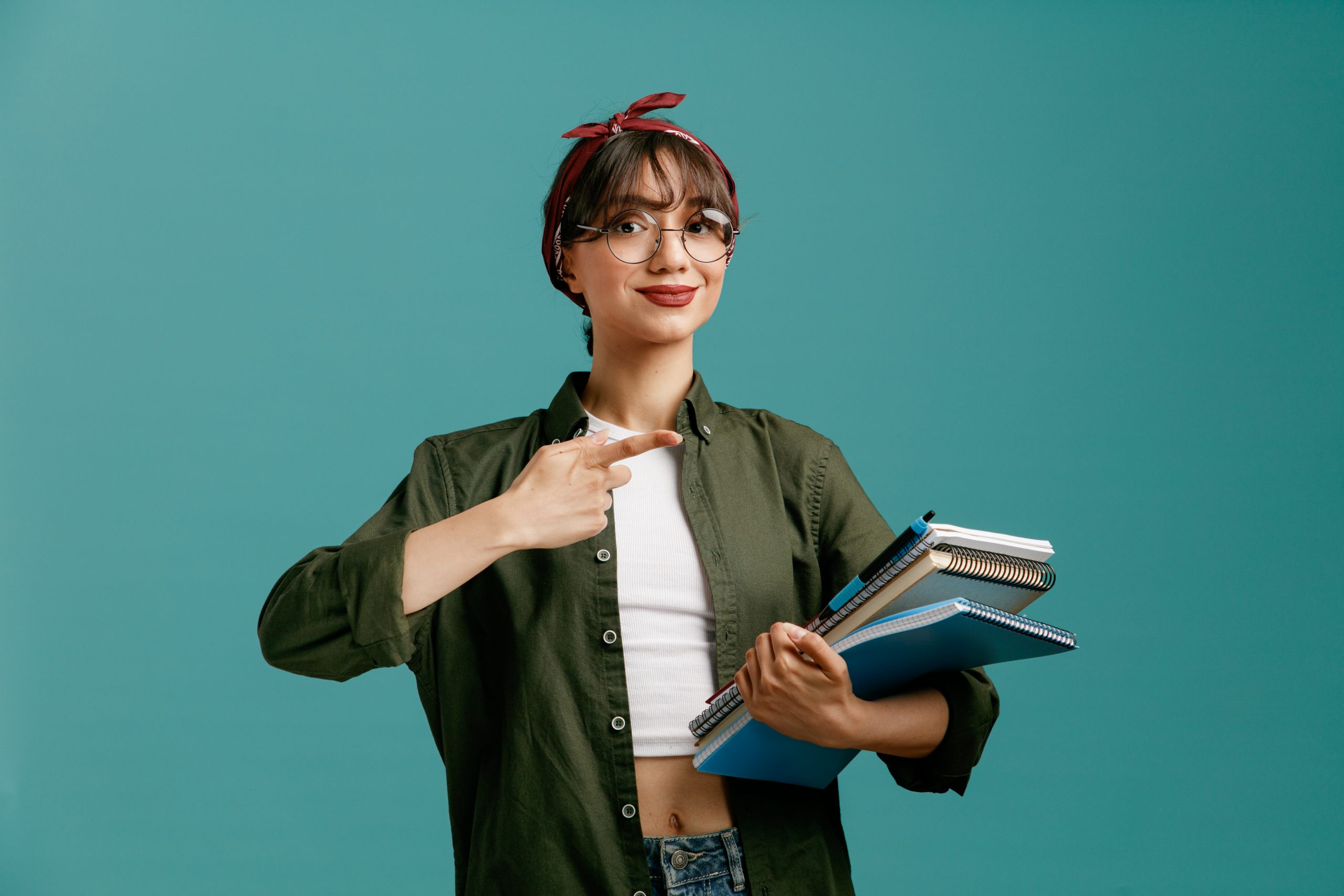 pleased young student girl wearing bandana glasses holding large note pads with pen looking at camera pointing at note pads isolated on blue background